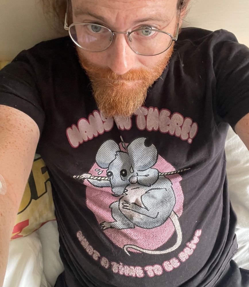 Picture of me wearing the What a Time to be Alive podcast shirt featuring a rat hanging from a rope that says “hang in there!”