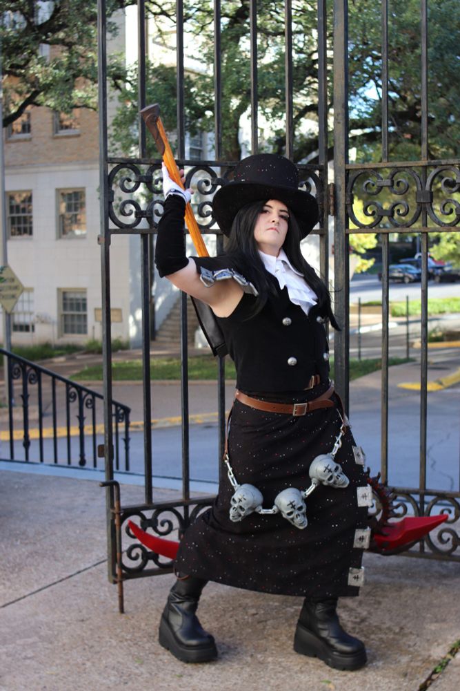 A cosplayer dressed as Testament from Guilty Gear Strive. His arm is raised, holding the handle of a large prop scythe that peeks out behind his calf. He wears black knee boots, a long black skirt with metallic details on the slit, a brown leather belt with three grey skulls hanging down from it, a black velvet vest with silver buttons, a single black velvet sleeve, a white glove, a white jabot with a stiff collar, long black wig, and a top hat.
