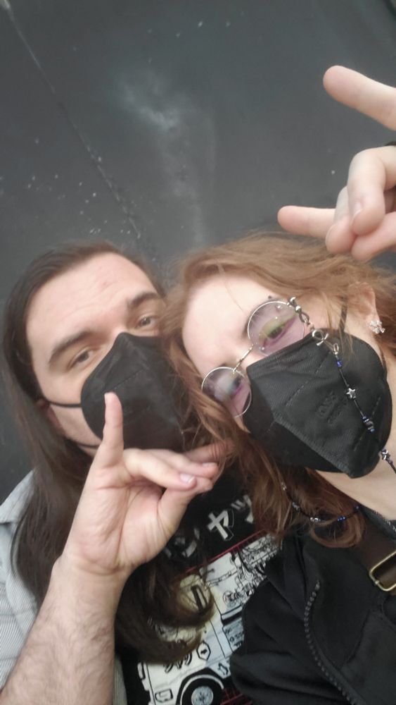 A selfie of two white people wearing masks holding up the Too Sweet hand sign