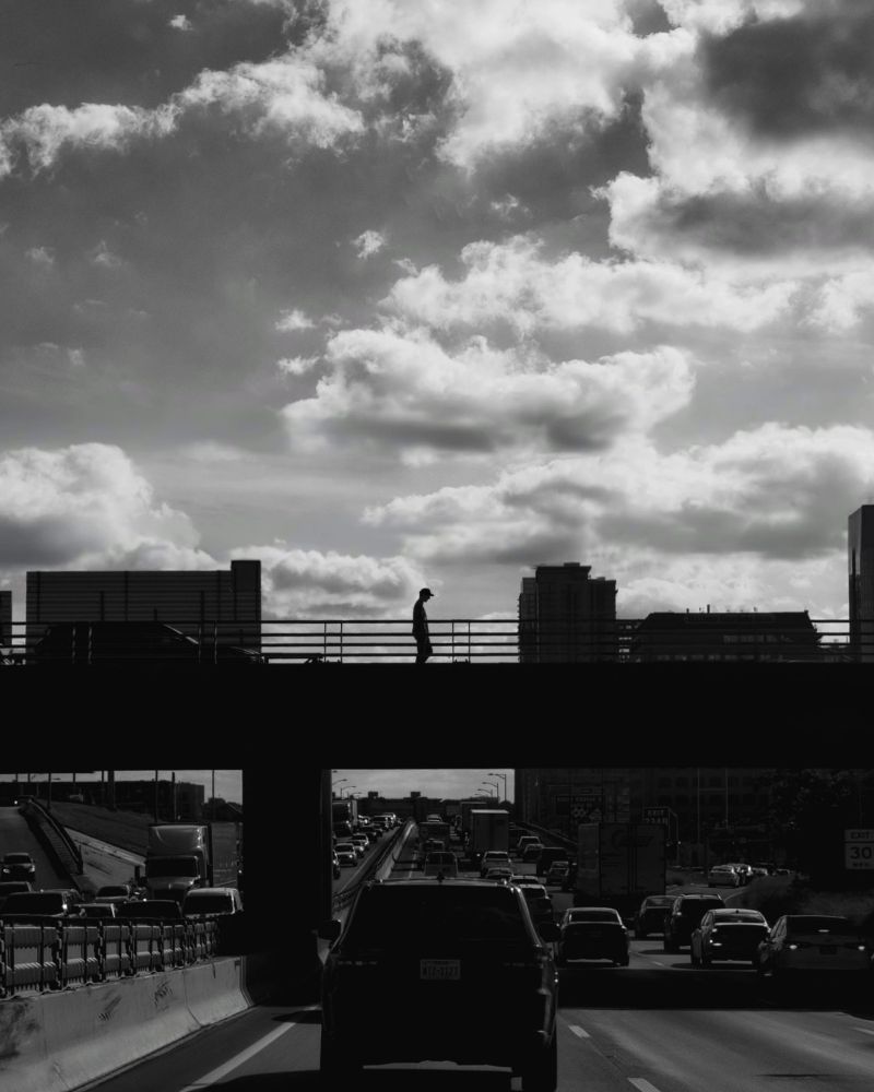 Sitting in traffic capturing a moment in time of a person walking across the overpass and cars lined up in both directions on the highway. Black and white street photography edition.. Rush Hour.