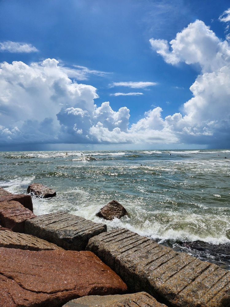 A photo taken from the Jetti of a bunch of cool rocks with the ocean breaking against them. The clouds are omnious with rain storms coming from a few of them, and if you look closely, you can see some surfers out in the water.