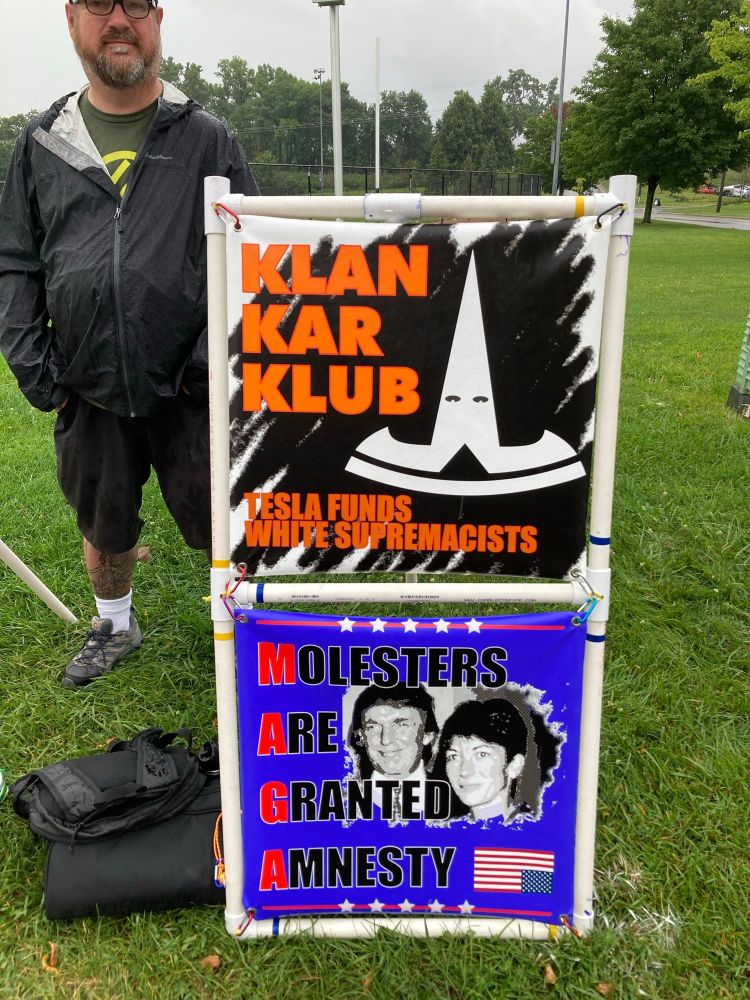 a man standing behind a pair of banners in a field; the first banner shows an inverted Tesla "T" emblem upside down, with eyes superimposed on the upper part, resembling a Klan hood; the text reads "Klan Kar Klub" and "Tesla Funds White Supremacists"

The second banner is an image of Donald Trump with Ghislane Maxwell, convicted of child sexual abuse as an accomplice to Jeffrey Epstein. The picture is on a blue background, with stars at the top and bottom, and an inverted American flag in the lower right corner. The text on the banner reads:
"Molesters
Are
Granted
Amnesty"
With the first letter of each line in bright red to spell out "MAGA". 