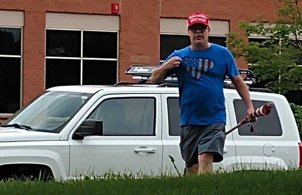 White male wearing a red "Make America Great Again" ball cap and a blue t-shirt with an American flag inside an outline similar to a Superman logo, pointing at himself with one hand while he holds a wooden flagpole and American flag in the other. He is standing in front of a white four-door SUV, possibly a Jeep. 

Taken at the Tesla Takedown protest in Golden Valley, MN on July 5, 2025, a few minutes after he marched from his car over to two older female protesters and started yelling at them and swinging his flagpole within an inch of their faces to try to intimidate them, only stopping when others intervened. 

He then retreated to his car, continuing to scream profanity at the protestors. 

He seems like a charmer. I wonder if any women in his life trust him? 