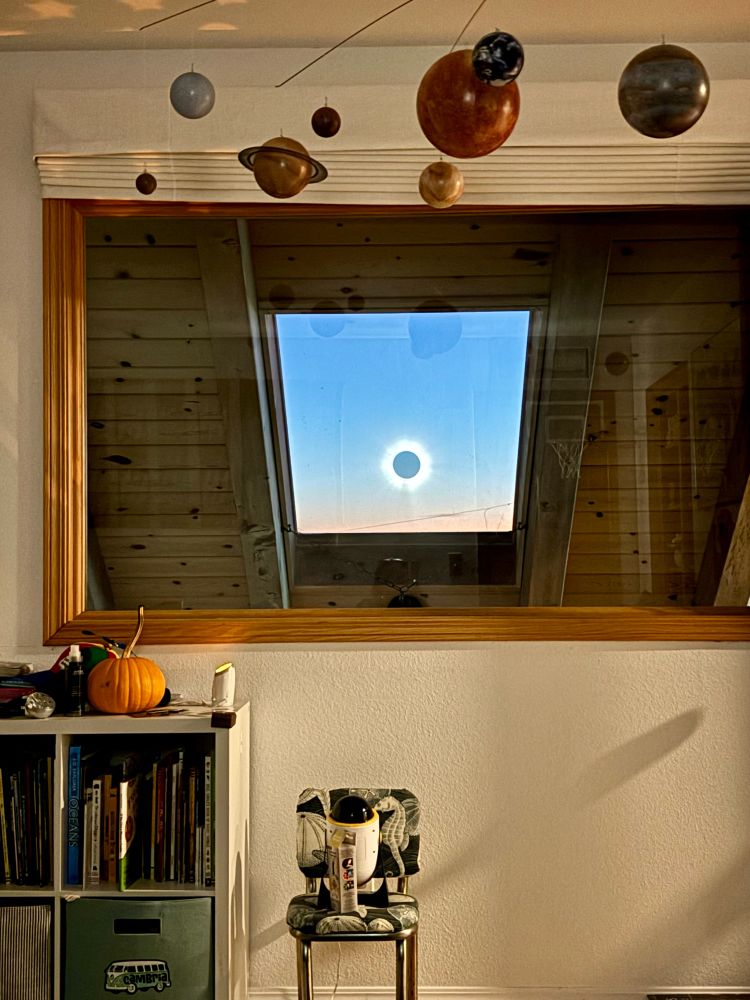 Kids room, view focused on a window and skylight, with a reflection that looks like a total solar eclipse. A solar system mobile hangs from the ceiling, and a small chair is centered under the window with a bookshelf to the left. 