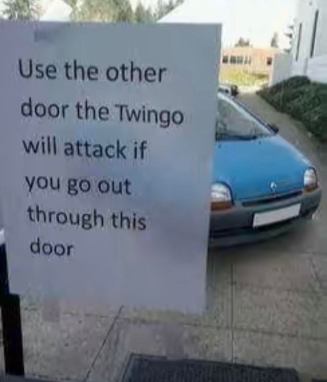 Low quality photo of a blue original twingo through a glass door. Only the front is visible behind a paper sign taped on the door saying ‘Use the other door the Twingo will attack if you go out through this door’