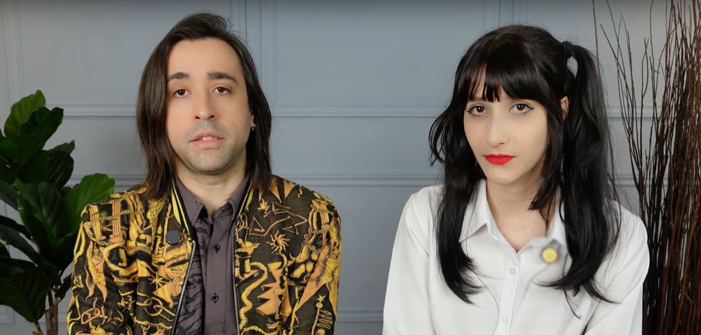 pedro medeiros and isadora sophia from coldblood inc. they are sat together in a room with plants behind them. pedros is wearing a black and gold blazer and isadora is wearing a white blouse and has blakc hair in pigtails.
