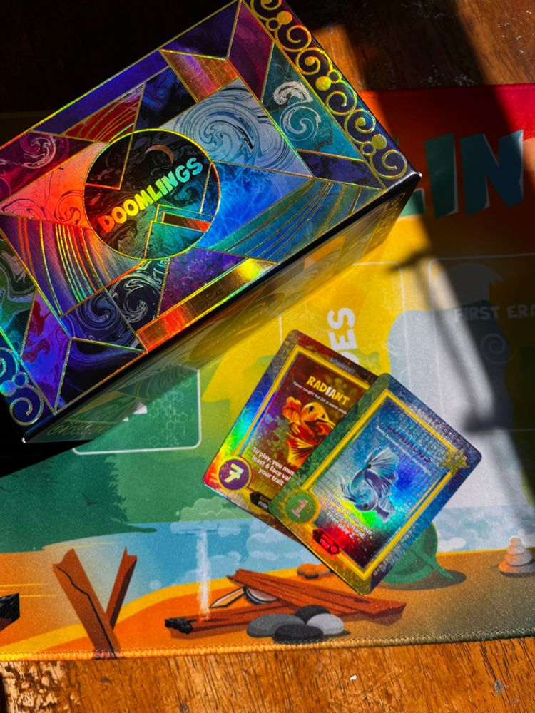 The two limited edition holofoil cards "Radiant" and "Luminous" posed next to the Doomlings Deluxe edition holographic box, set atop a Doomlings Playmat, all glittering/shining in the sunlight.