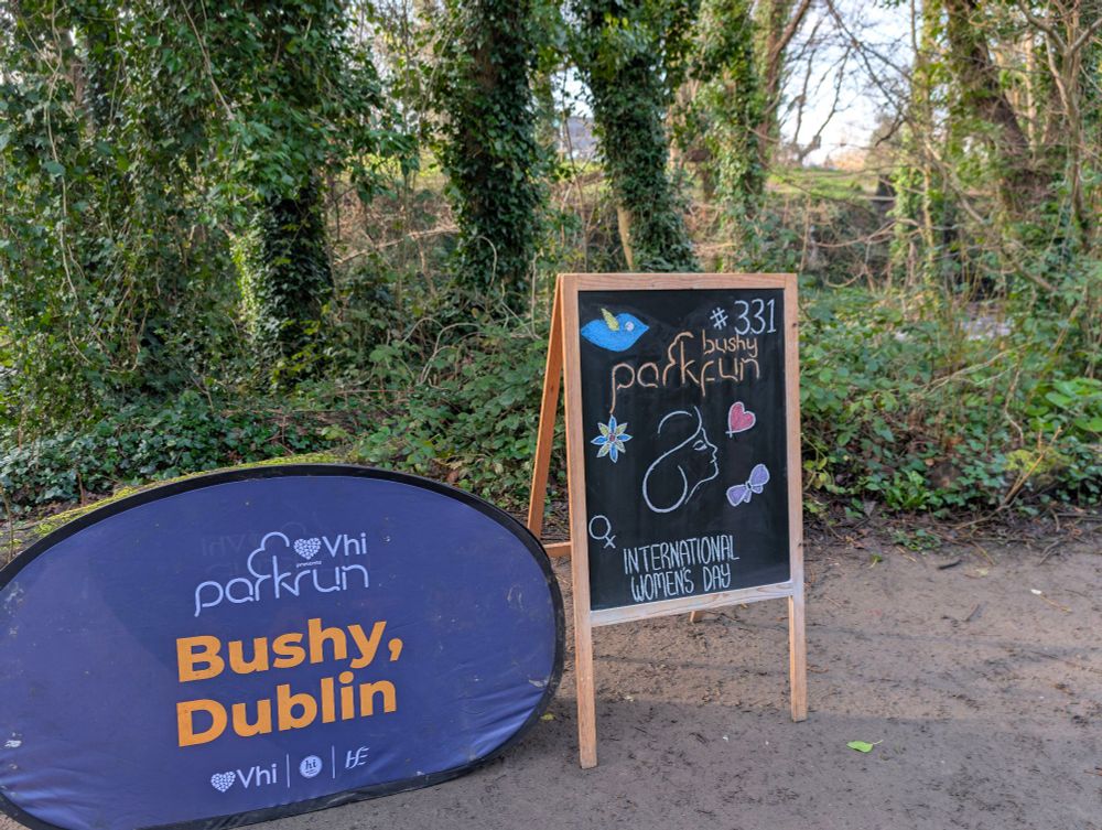 Popup sign and sandwich board on muddy ground with trees and bushes beyond. The sign reads Parkrun Bushy Dublin.