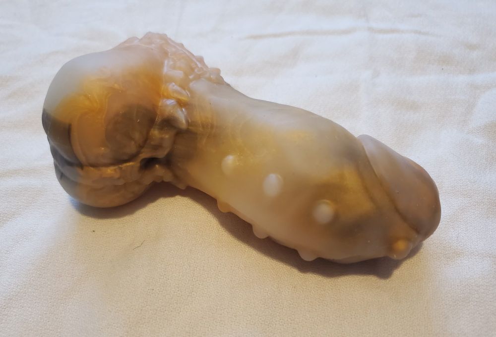 A gold, black and unpigmented Throgul from Made to Were. Throgul is a short and girthy phallus with piercings and a swell. The faux-harness around the balls makes an excellent spot to grind against.