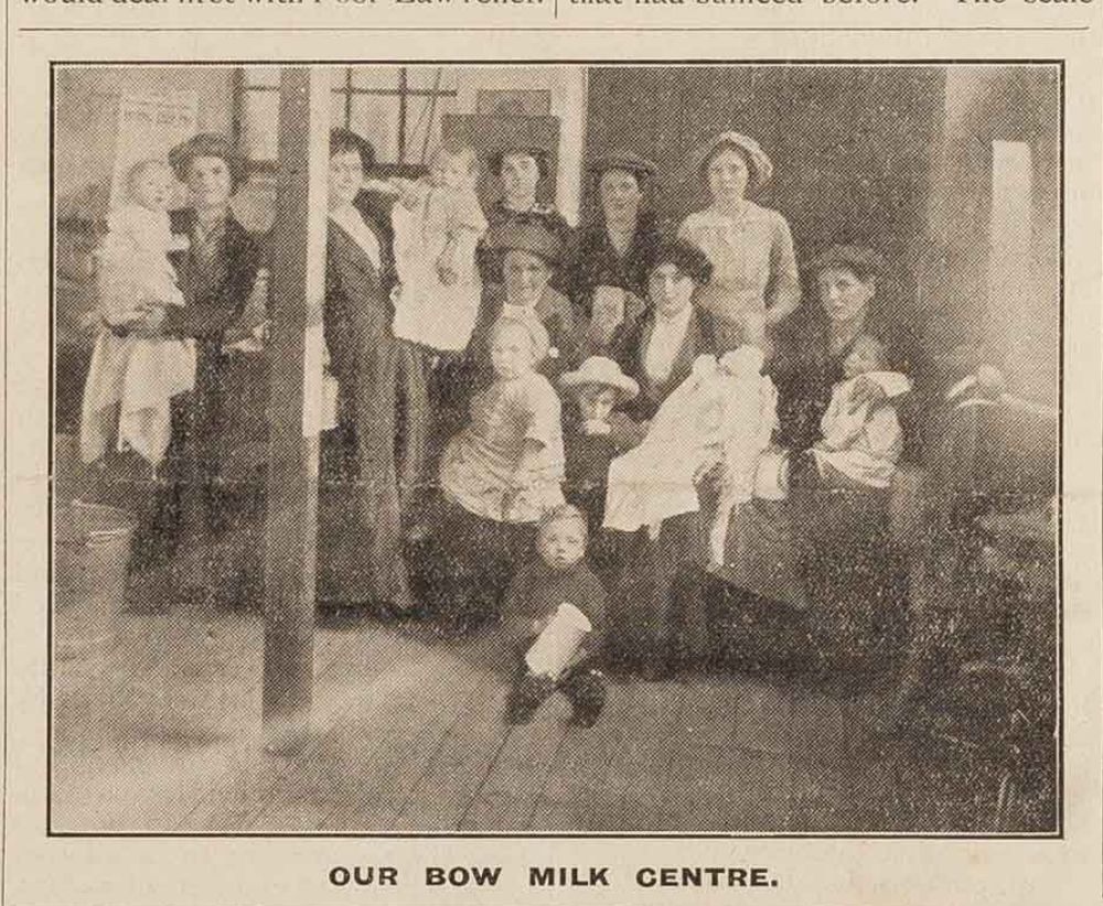 An image from the cover of the Sept 26, 1914 issue of Sylvia Pankhurst's paper, the "Woman's Dreadnought," shows several women, most of them holding small children, in a wood-floored room in the East London neighborhood of Bow. An especially chonky little boy sits in the foreground, looking at the camera and holding a milk pail. The caption reads "Our Bow Milk Centre." 