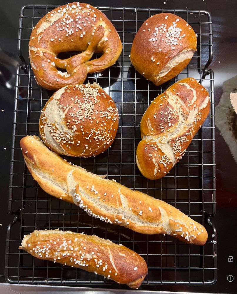 1 schwäbische Laugenbrezel (erkennbar am charakteristischen Einschnitt und dem prägnanten Bauch – hier eher als halb misslungener aber sympathischer Versuch erahnbar), 2 Laugenbrötchen und 3 unterschiedlich geformte Laugenstangen auf einem Gitter, dass auf einem Ceranfeld steht. Die Teiglinge wurden vor dem Backen unterschiedlich bestreut. Teils mit grobem Salz, teils mit Sesam.