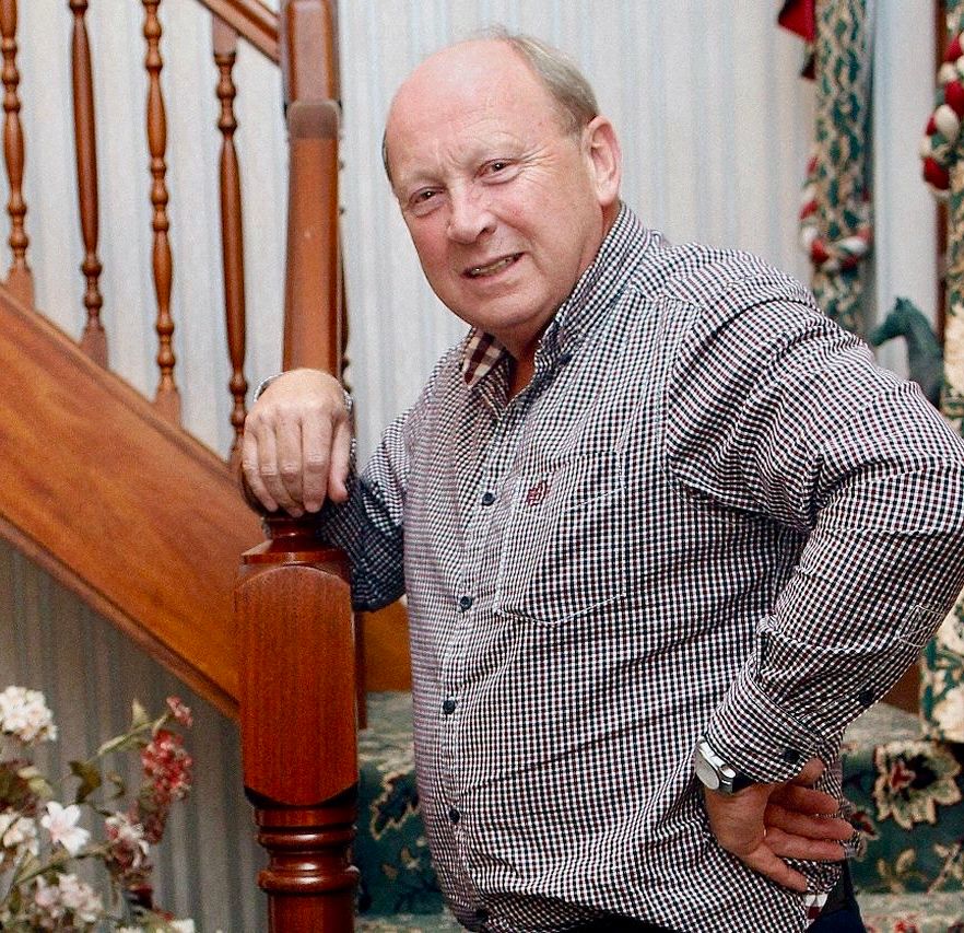 The TUV’s Jim Allister standing at the bottom of a staircase in a traditionally decorated home (of course, no modern or foreign influences tyvm) wearing a red white and blue check casual shirt (of course, no explanation necessary), one hand on hip, the other coquettishly draped over the newel post, and giving his best come to bed eyes.