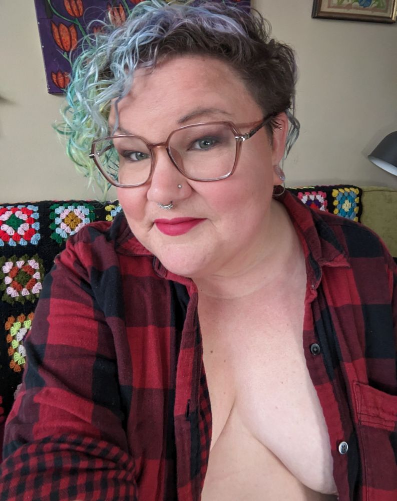 Greer is wearing an unbuttoned red buffalo plaid flannel shirt. She sits on her couch while smiling at the camera with big red lips. She is wearing large glasses and her nostrils and septum are pierced. 