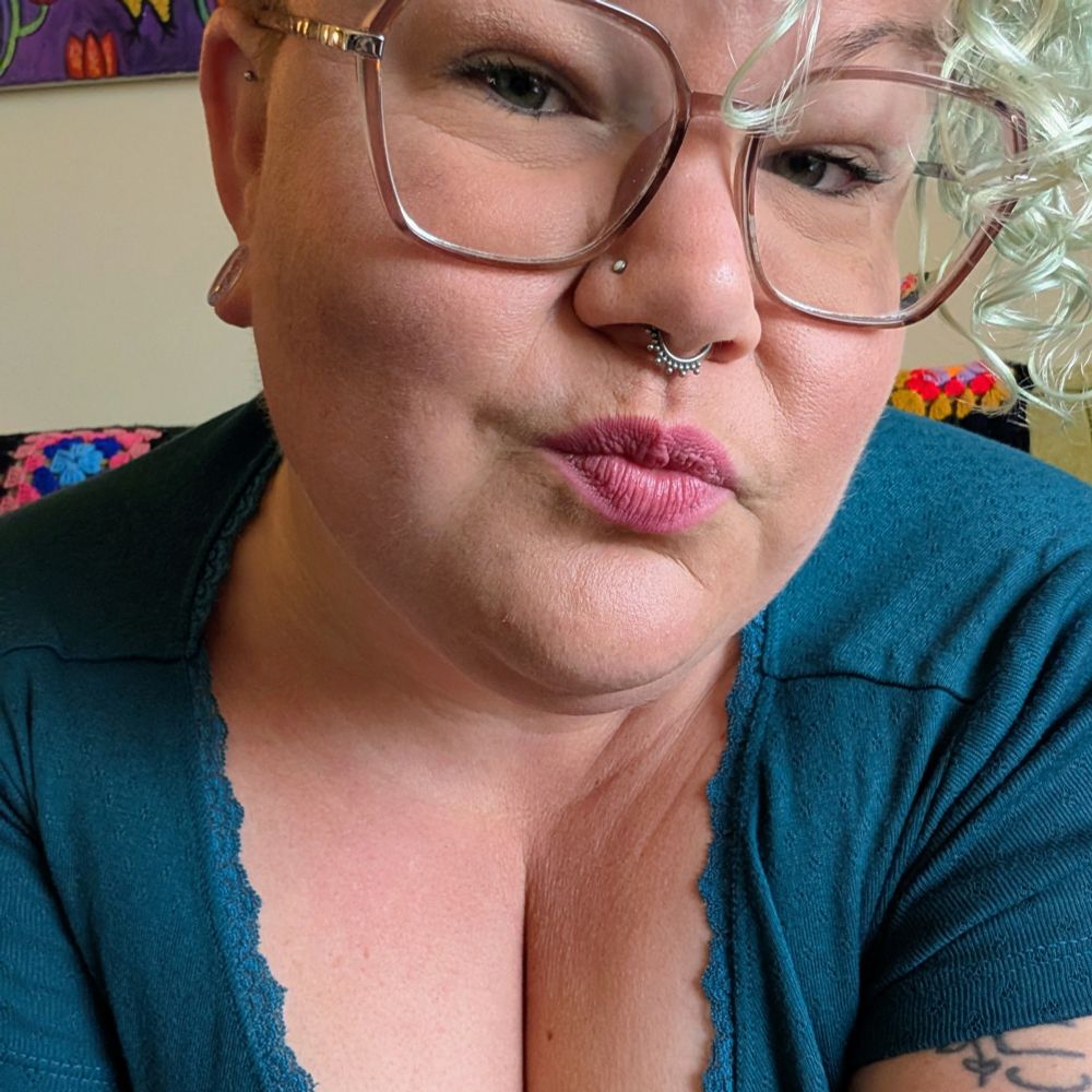 Greer smiles at the camera. She is wearing a low cut teal shirt and glasses. 