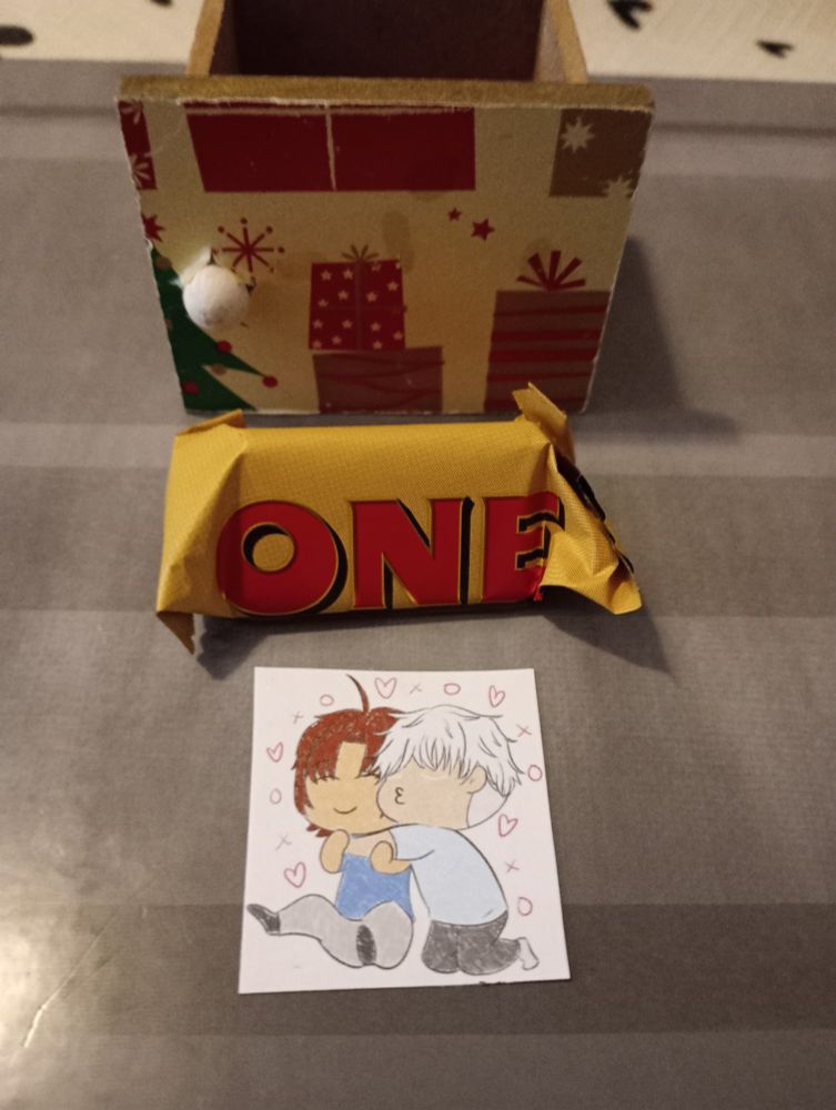Image shows a small drawer with part of a christmas tree and some presents on it Infront of which is a mini toblerone and an artwork in chibi style that shows Odasaku from Bungo Stray Dogs smiling whilst being hugged and kissed on the cheek by Gojo Satoru from Jujitsu Kaisen they are in casual blue and grey clothes 
