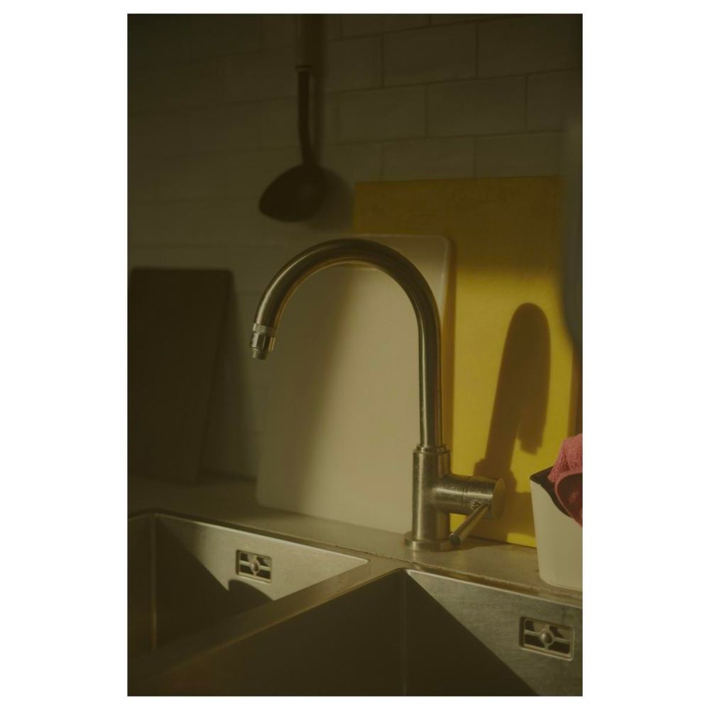 Zwei Edelstahlwaschbecken nebeneinander. Dahinter zentral ein rund gebogener Wasserhahn. An der weiß gefliesten Wand rückseitig stehen versetzt zwei Brettchen. Eins weiß, das andere gelb. Die Szene liegt im dunklen diffusen Licht, abgesehen von einem Quadrat aus Morgensonne, welches den Wasserhahn und die Brettchen in weiches Licht taucht. 