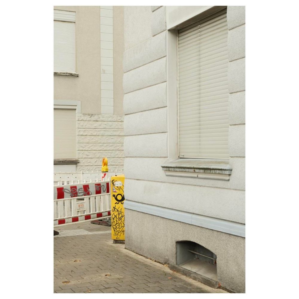 Eine Straßenecke. Die Häuser sind weiß-grau. Vor den Fenstern sind bei allen Häusern die Rolladen herabgelassen. Auf der Straße steht eine rot-weiße Straßenabsperrung neben einem gelben Briefkasten. 