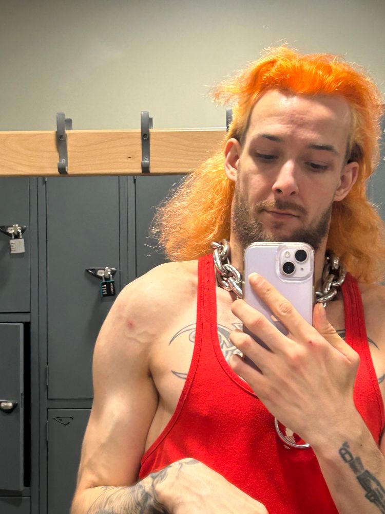 Me flexing my right tricep and seeing a lil vein appear. I have an orange mullet and a red tank top on. 