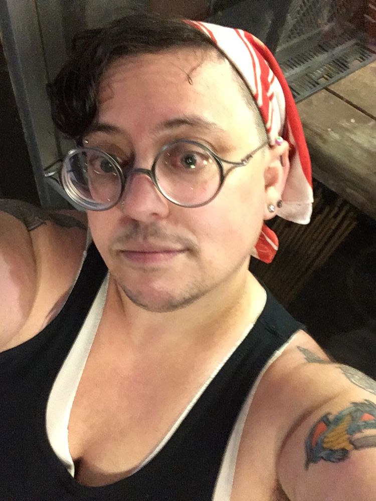 Selfie angle photo of a light-skinned person with round glasses, wearing a binder and black tanktop. They are wearing a red and black bandanna over short hair.