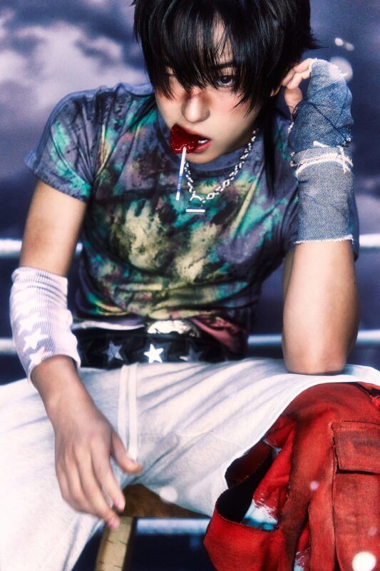 The same man as the previous photo. He sits his his legs spread, and the camera looks up from between them. He's made up to look like he's been fighting, with wraps on his arms, and bruises on his face. He looks up from under his bangs with a red lollipop dangling sexily from his lips.