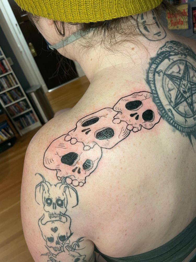 (Close up of the skulls) The back of a client, showing a fresh tattoo of three cartoon demon heads stacked on top of each other on the right shoulder, and three cartoon skulls stacked on top of each other on the left shoulder, both oriented towards an older pentagram tattoo that was not done by me.