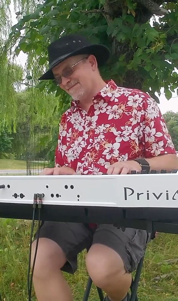 A pianist in a black hat and bright, flowery, red shirt plays a digital keyboard outdoors.