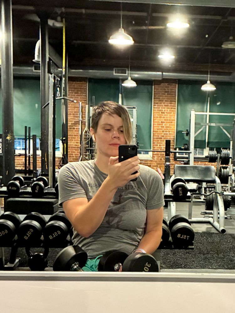 White latine queer in the free weight area of a community gym. They are looking down at their phone with a slight smirk and wearing a gray tight top with dumbells on their lap. There's long hanging lights and exposed brick in the background.
