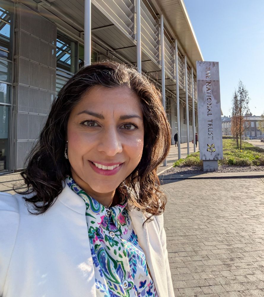 Woman smiling at camera standing in front of a building which is the headquarters of the National Trust. She is wearing a floral shirt and jacket.