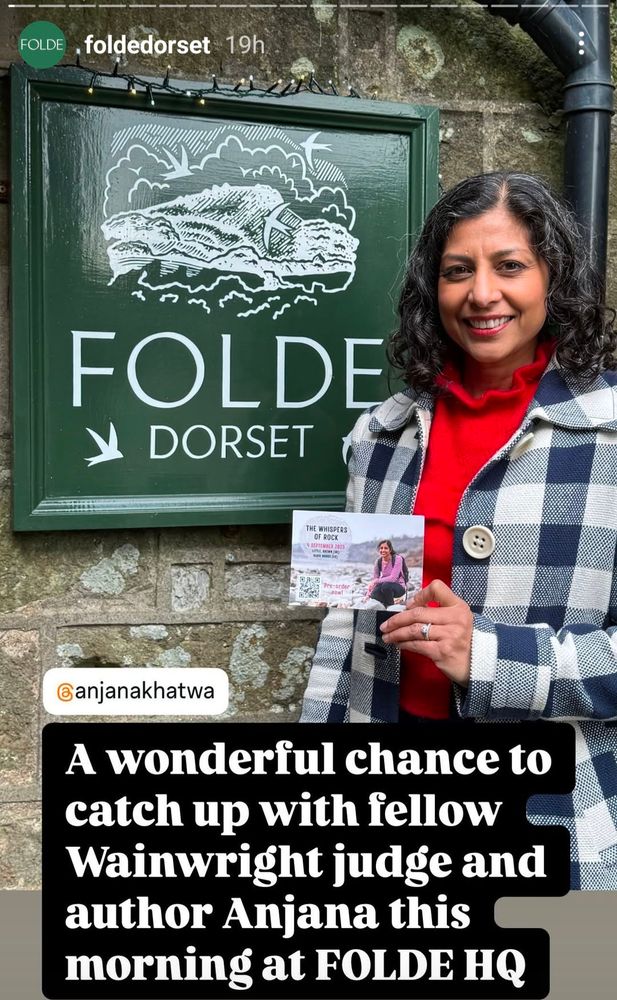 A woman wearing a blue and white gingham coat with a red sweater beneath. She is standing in front of a green shop sign that says Folde Dorset. She is holding a postcard that advertised her forthcoming book The Whispers of Rock. 