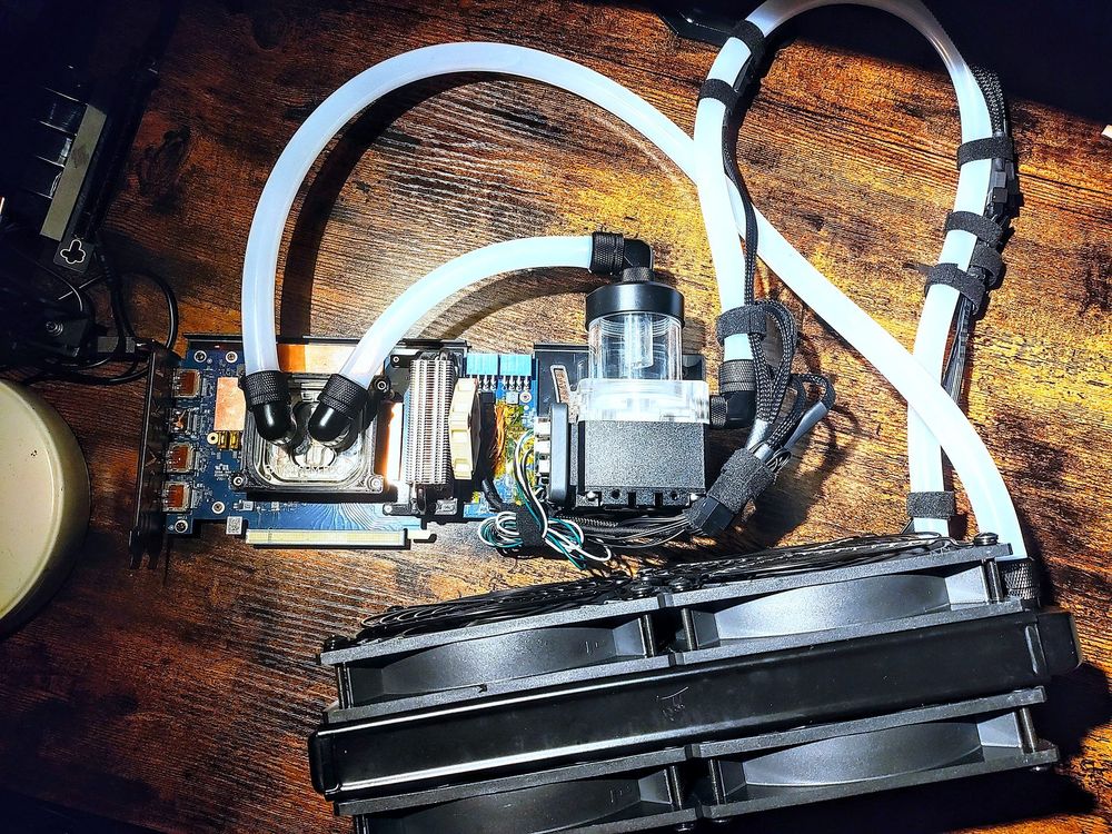 an Arc A770 graphics card attached to a liquid cooling loop sitting on a wood tabletop. White silicone tubing connects the waterblock, pump, and a radiator with push+pull fans.