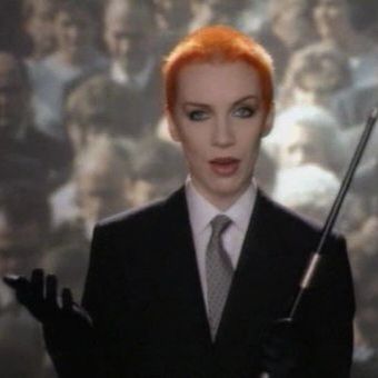 Androgynous with short bright orange hair in black suit. White dress shirt and gray tie underneath.  Behind is large image of crowded sidewalk.