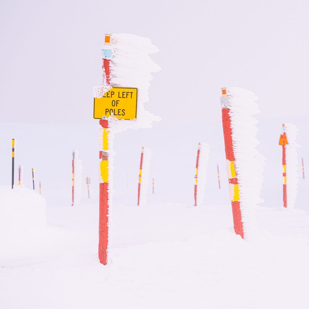 Keep Left depicts bright red and yellow roadside markers at Mount Hotham, Australia. The snow clouds are a similar tone to the ground, creating a slightly surreal scene where many poles of various sizes are staggered against each other. The poles have very directional ice build up of about two feet, reflecting the prevailing wind direction and the cold conditions.