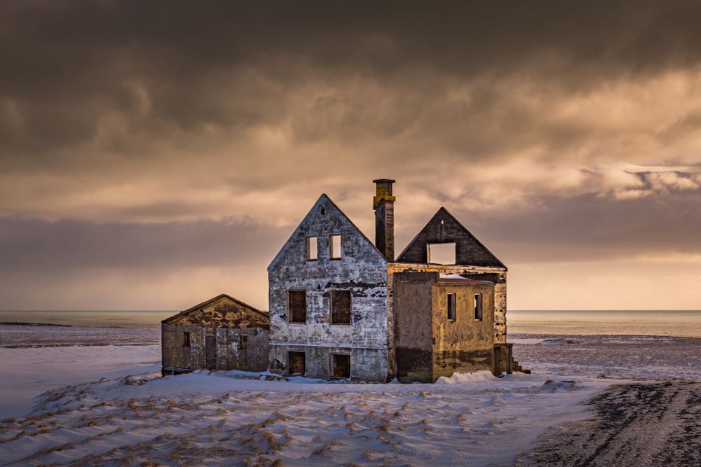 An old abandoned Icelandic farmstead missing its roof sits in snow near the coastline of Iceland. Above a distant snowstorm is illuminated in golden tones from the low afternoon light.