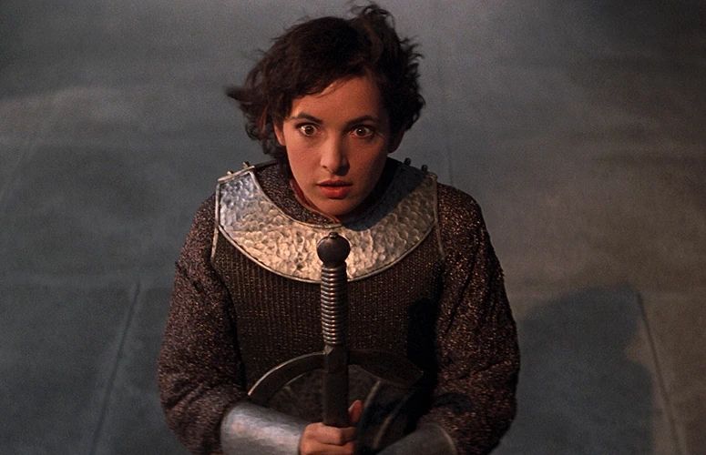 Jane Wiedlin as Joan of Arc, wearing French armor, kneeling, looking at the camera, holding a sword downwards in front of her. From Bill & Ted's Excellent Adventure.