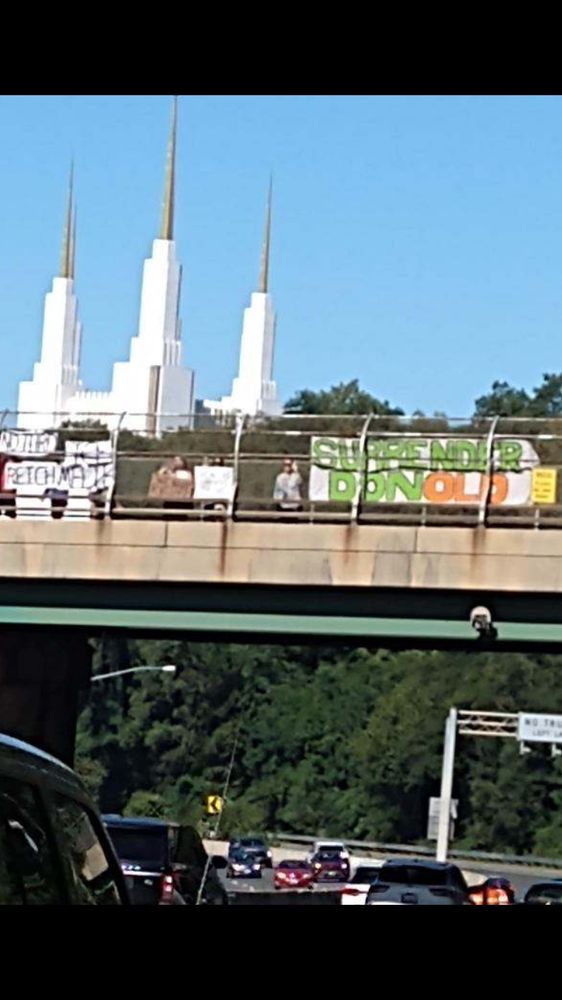 Banner sign on highway overpass that says Surrender DonOLD- in the distance is the spires of the Mormon Temple. 
