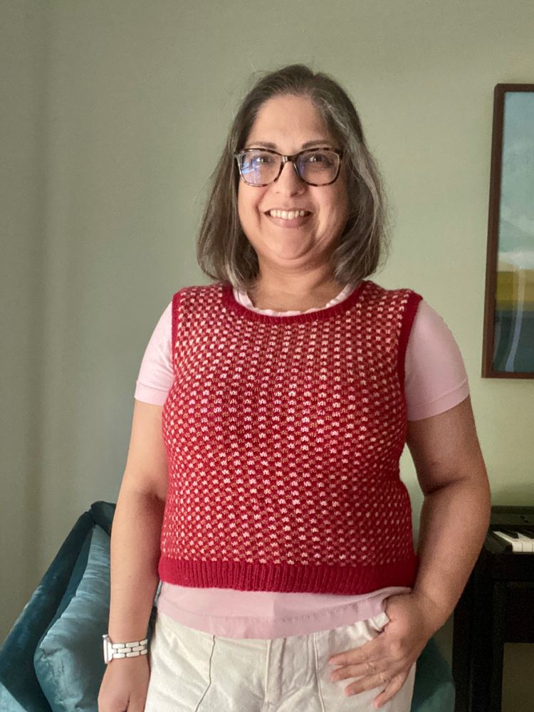 A middle-aged brown woman stands in front of a green wall in a red hand knit vest, pink t-shirt and cream pants.