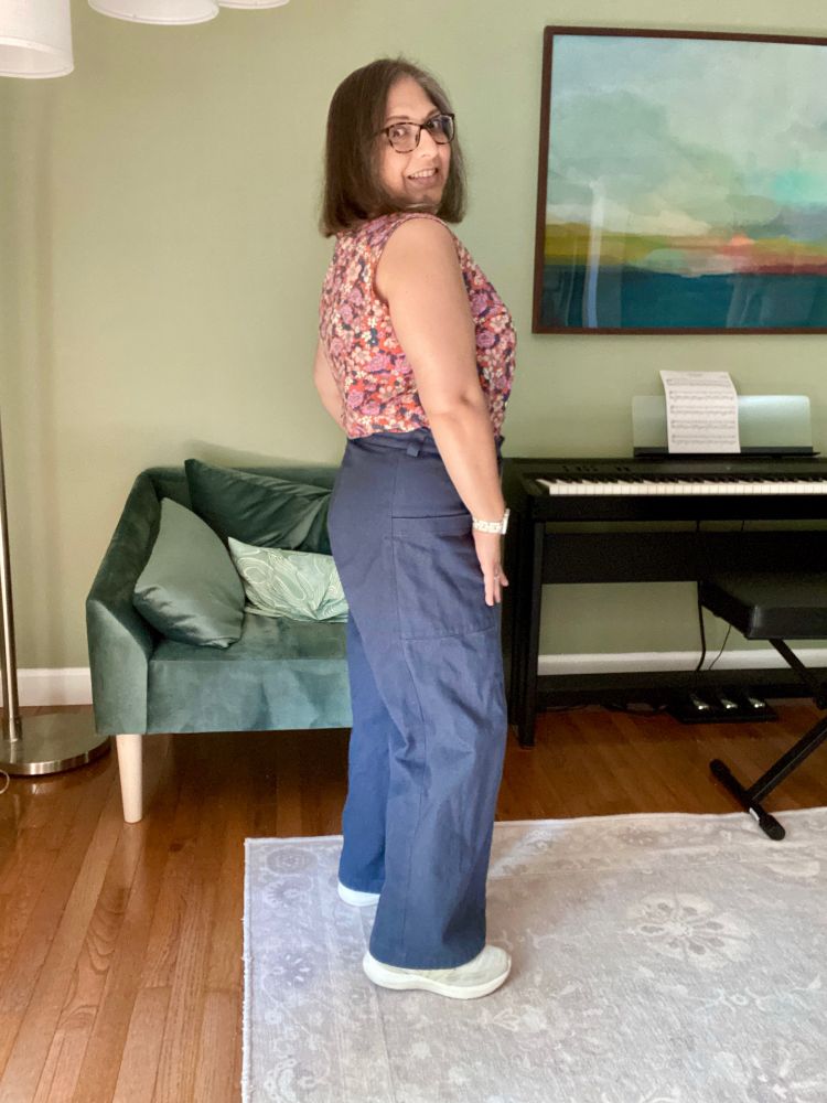 A short middle-aged brown woman stands facing sideways in blue pants and a floral tank.
