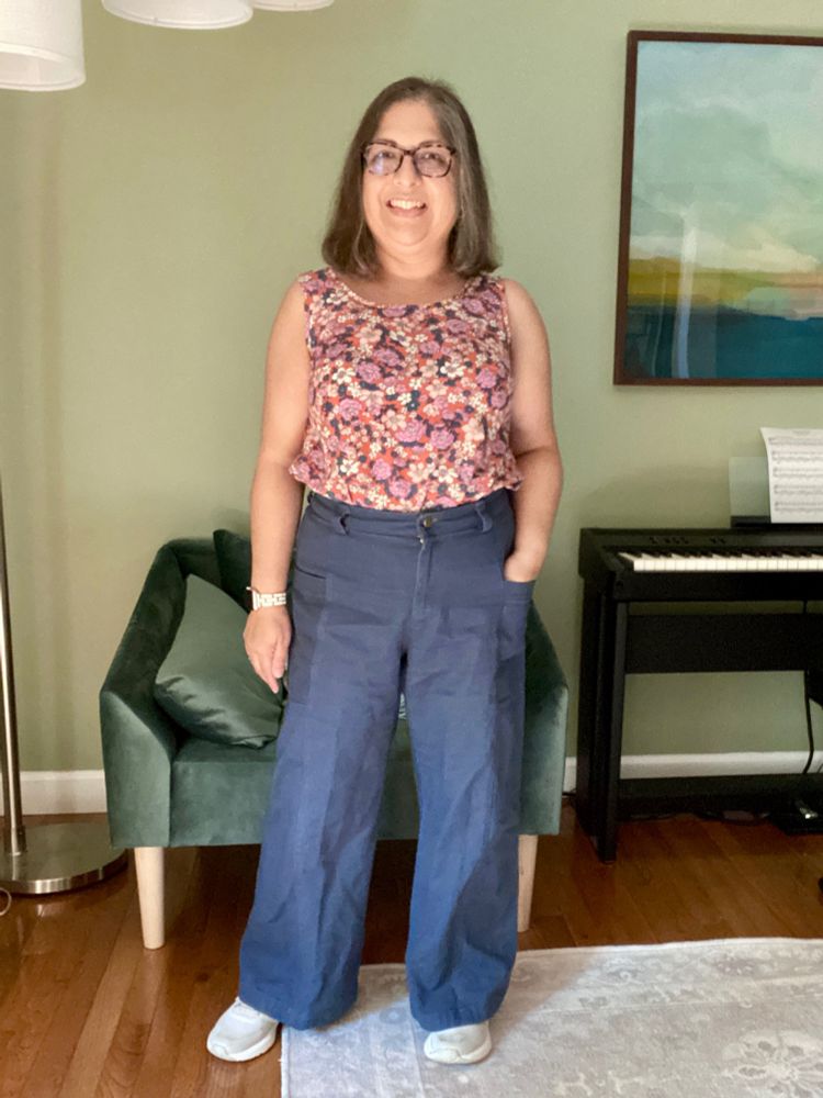 A short middle-aged brown woman stands with her hand in one pocket. She is wearing blue wide-legged pants and a floral tank with white sneakers.