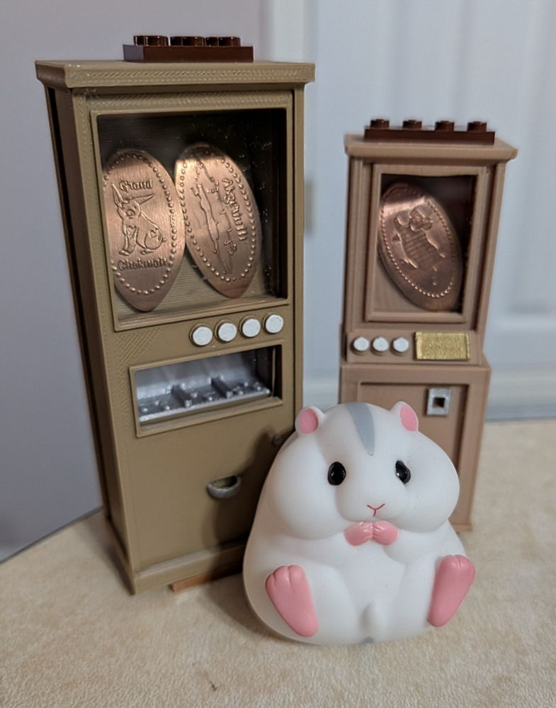 Charles the hamster (toy) sitting in front of two 3D printed mini pressed penny machines. The smaller machine has a hamster pressed penny in it, the larger has two coin designs related to the game Tales of the Abyss