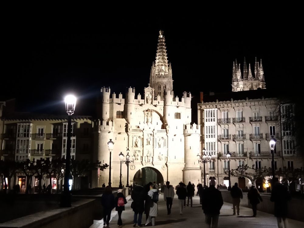 Vista nocturna de de la Catedral de #Burgos desde el puente de Santa María.  Se ve a gente en el puente, una vista completa del Arco de Santa María flanqueados por dos edificios de corte decimonónico.  Sobre este asoma la torre sur de la Catedral.  Más a al derecha sobresale el cimborrio sobre el tejado del edificio aledaño al Arco.  También se ven los árboles del Paseo del Espolón, a la derecha, y el Pasea de la Audiencia, a la izquierda.