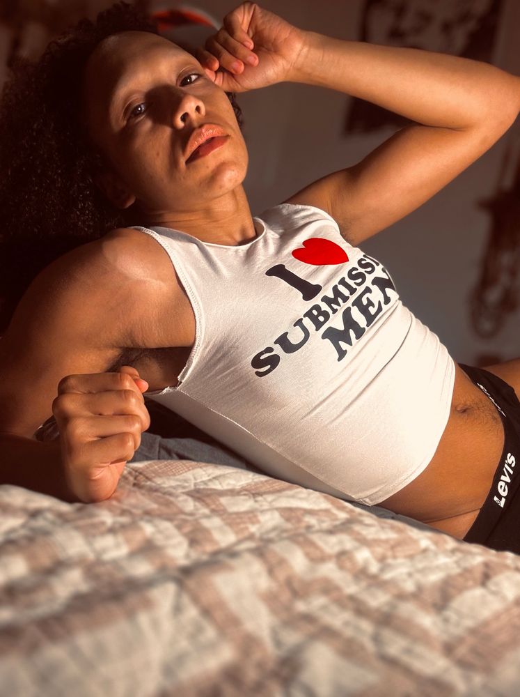 An androgynous person of African descent with light brown skin wears a white shirt that says “I love submissive men.” Their muscles bulge politely and you can see some of the veins under their skin. You can see a defined crease on their hip leading down to their underwear. They have long dark curly hair with a silver streak. They have hairy armpits and a happy trail for hair on their belly. In this image they’re lounging back on a bed, the camera low with them, focusing on their torso and face which is tilted upward, lips looking plump and inviting. They lean on one flexed arm and the other is bent upward gently touching their temple.