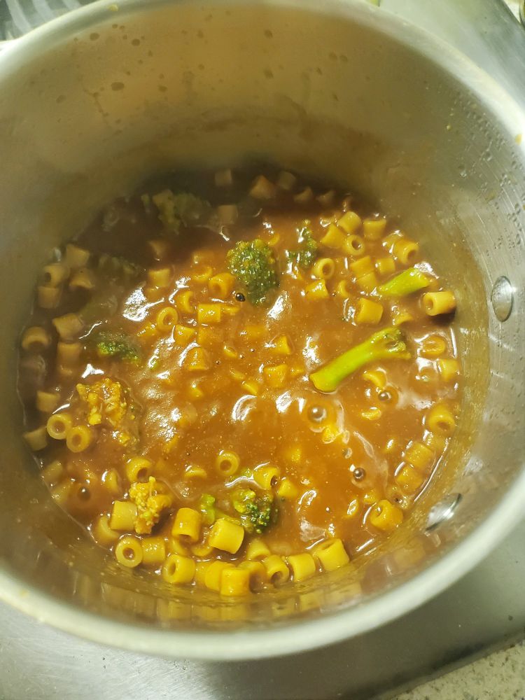 A stainless-steel pot filled with short, tube-shaped pasta and sauce, with some broccoli florets tossed in at the end for health reasons