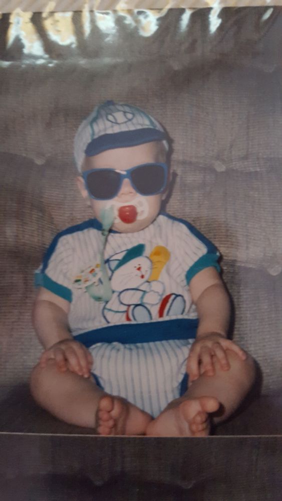 Baby Ryan sitting against a couch cushion wearing a hat, shirt, and shorts striped to look like a baseball uniform. He has a pacifier in his mouth, and sunglasses that make him look extremely chill as he looks straight into the camera.