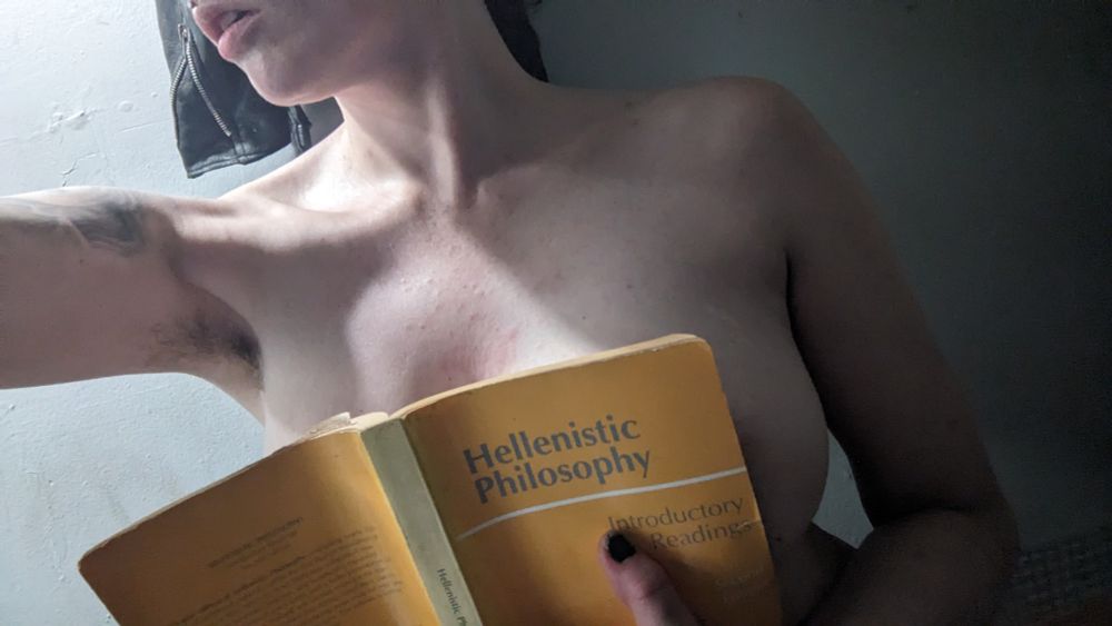 A picture of Lysi naked from the waist up w the top half of her face out of frame &her head turned to the side displaying her neck &collarbones. She is holding a mustard yellow book of Hellenistic Philosophy to her chest w her right sideboob uncovered &w just a tiny sliver of areola visible. One arm is outstretched showing off her luscious armpit hair.
