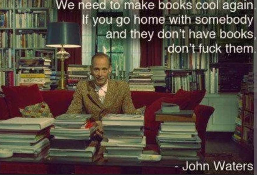 A picture of John Waters sitting on a red couch in a study surrounded by bookshelves &behind a coffee table covered in stacks of books. There are a wonderfully ridiculous number of books in said study, they are literally everywhere. Waters looks dapper as always &is wearing a light brown suit. Superimposed text says "we need to make books cool again. If you go home w somebody &they don't have books, don't fuck them"