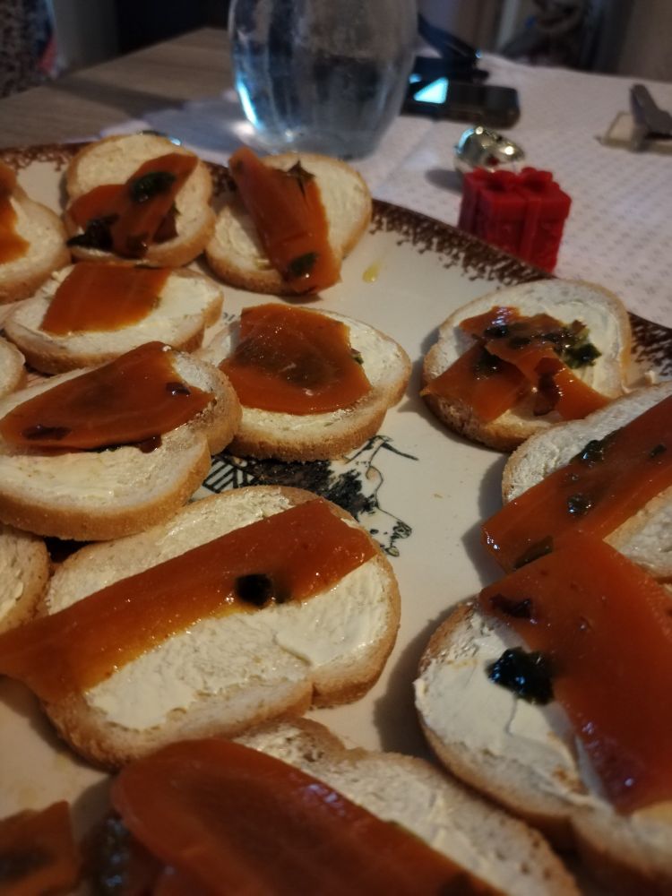 Des toasts beurrés au fauxmon sont dans une assiette blanche sur une table de fête. En arrière plan on y voit une bougie en forme de cadeau une cruche d'eau et un portable.