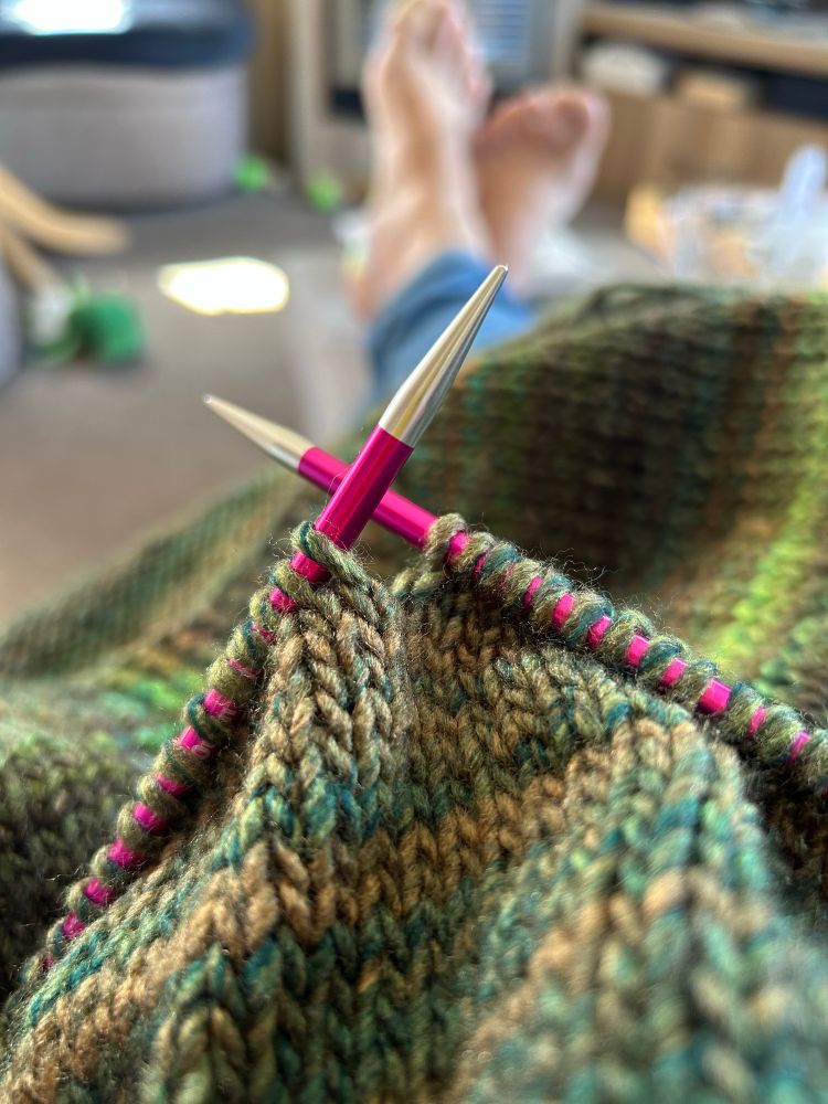 Knitting needles holding a piece of green and dark cream knitting. In the background I have my feet up
