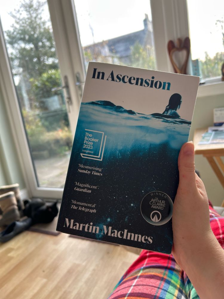 my hand is holding a copy of Martin MacInnes’ In Ascension. in the background is a glass door with a cat sitting in front of it 