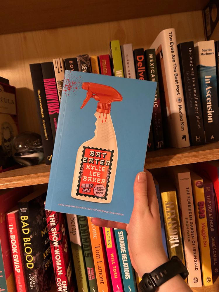 my hand is holding a copy of Kylie Lee Baker’s Bat Eater with bookshelves behind it