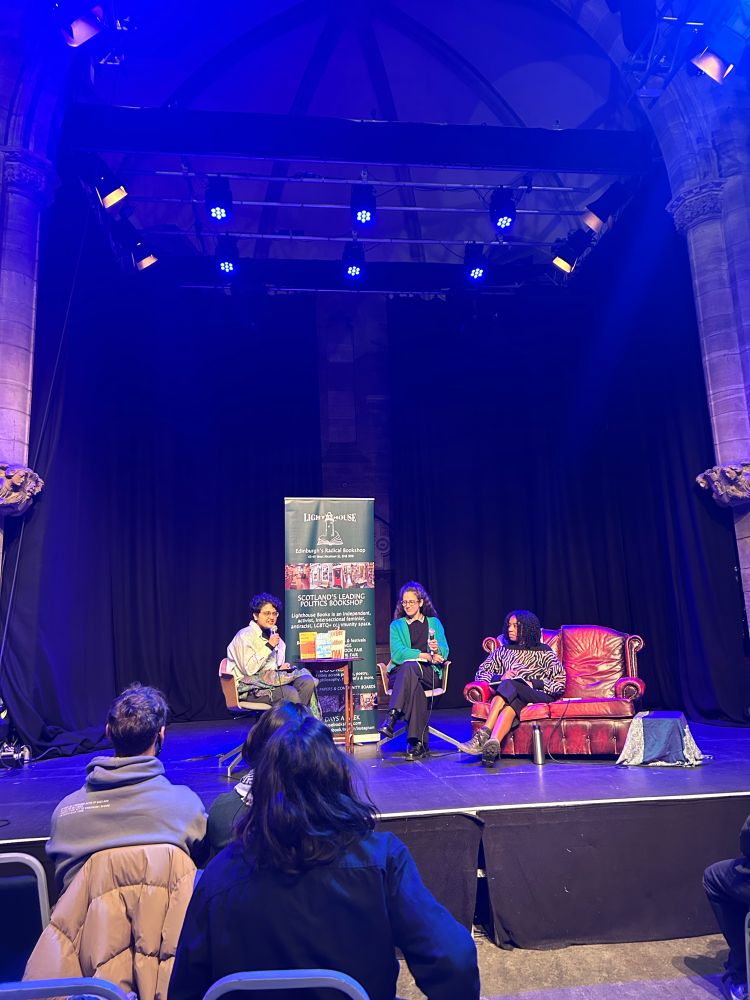 Nish Doshi, Leah Cowan, and Gracie Mae Bradley sit on a stage with a banner for Lighthouse Books behind them